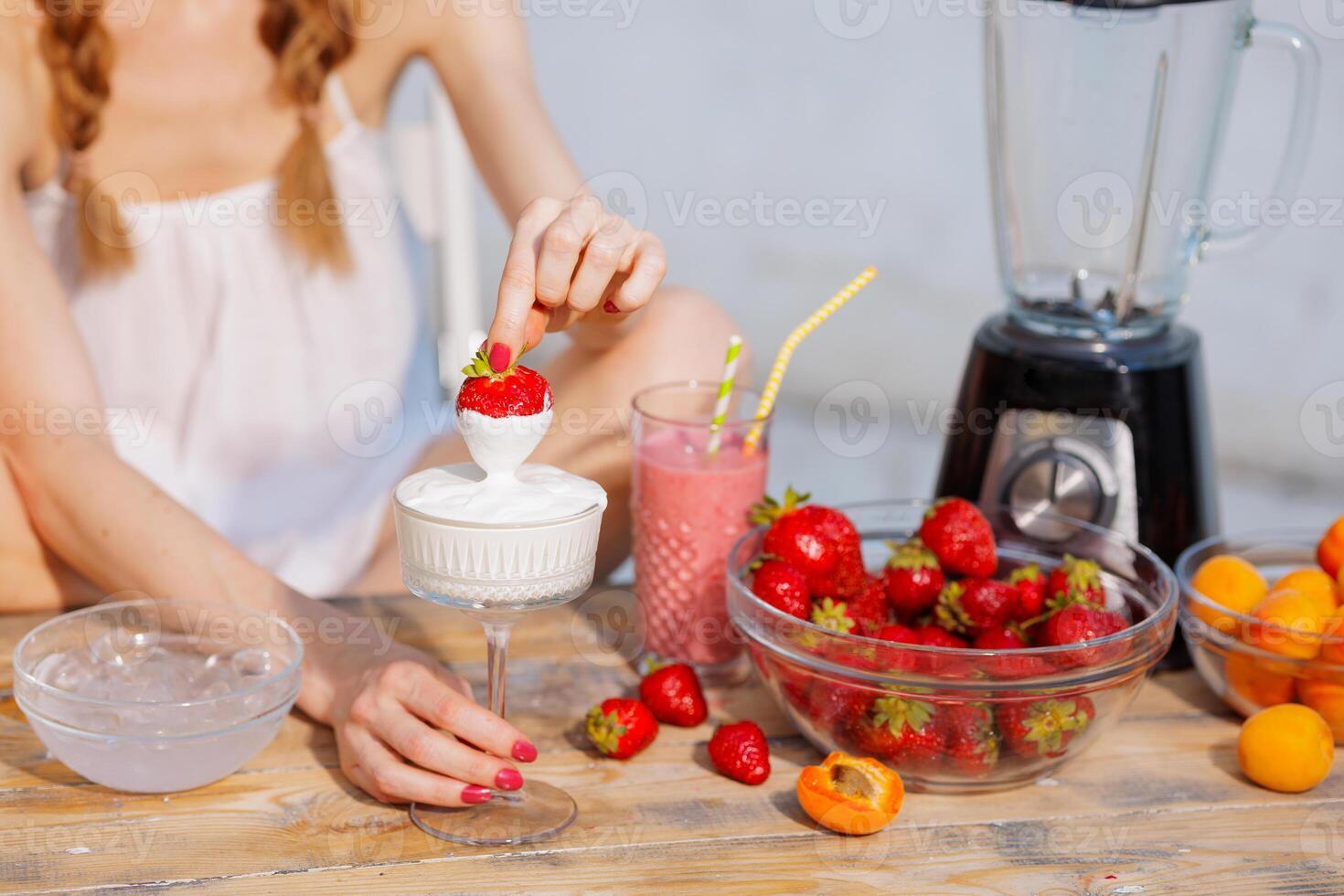 Close-up of a woman dipping a fresh strawberry into whipped cream at a rustic summer table with smoothie, blender, apricots, and strawberries. Fresh and sweet healthy dessert moment. photo