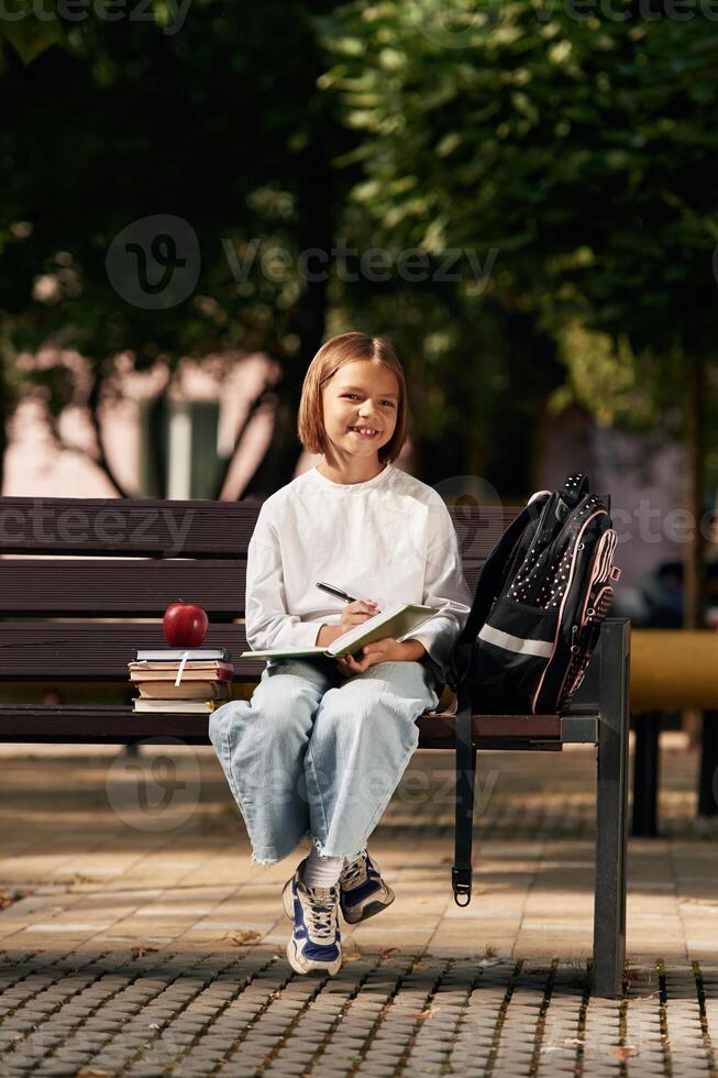 Writing ideas into notepad. Schoolgirl,,, is sitting on the bench with a bunch of books outdoors photo