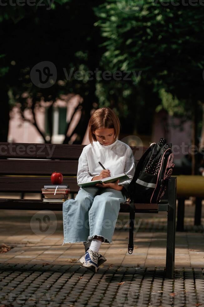 Writing ideas, into ,notepad. Schoolgirl is sitting on the bench with a bunch of books outdoors photo