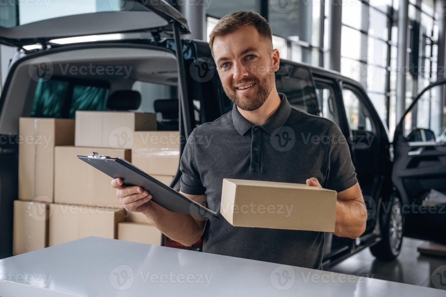 Positive facial expression, holding,, notepad. Delivery man or loader is near the car with a box photo