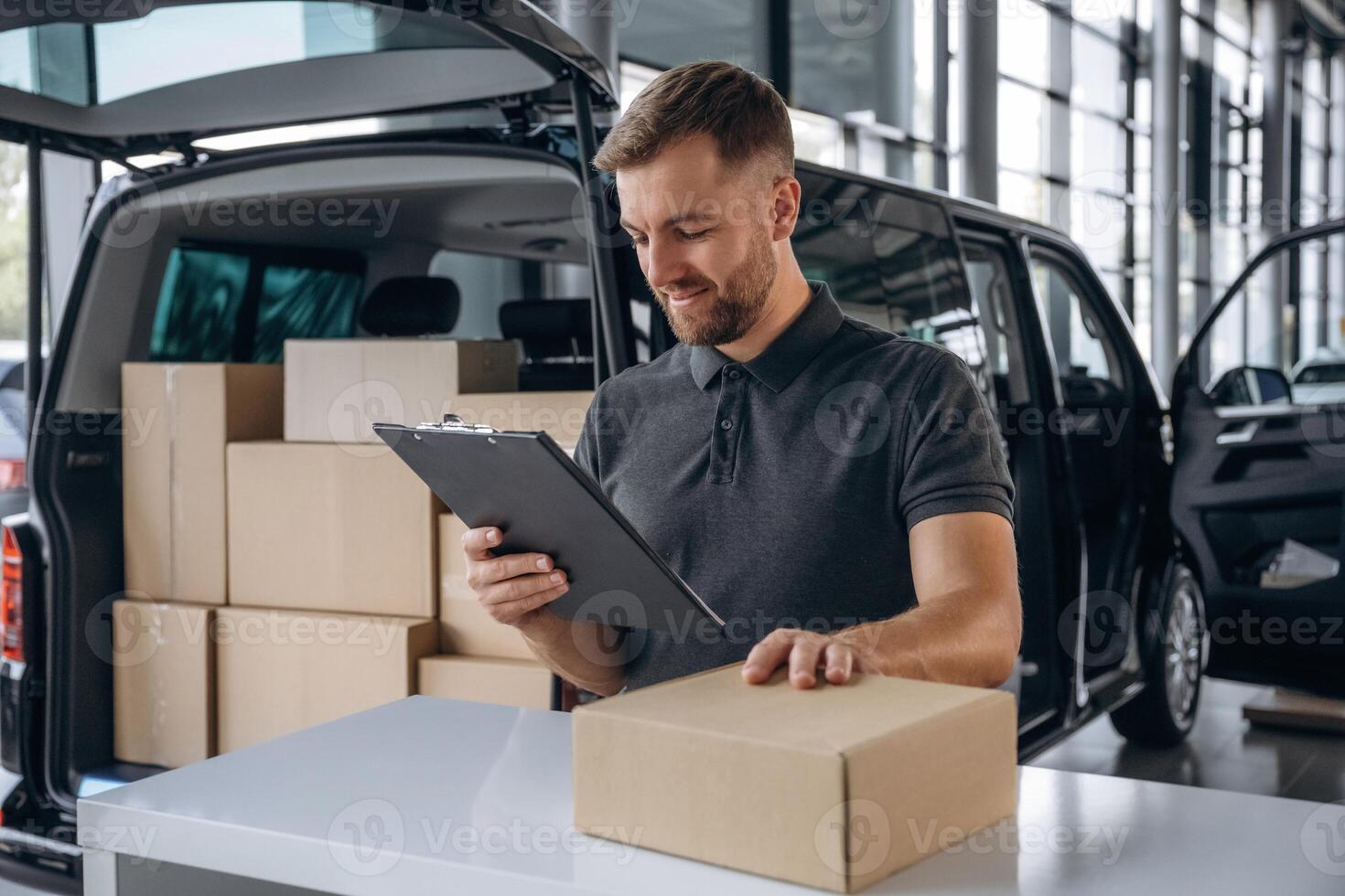 Positive facial expression, holding ,notepad. Delivery man or loader is near the car with a box photo