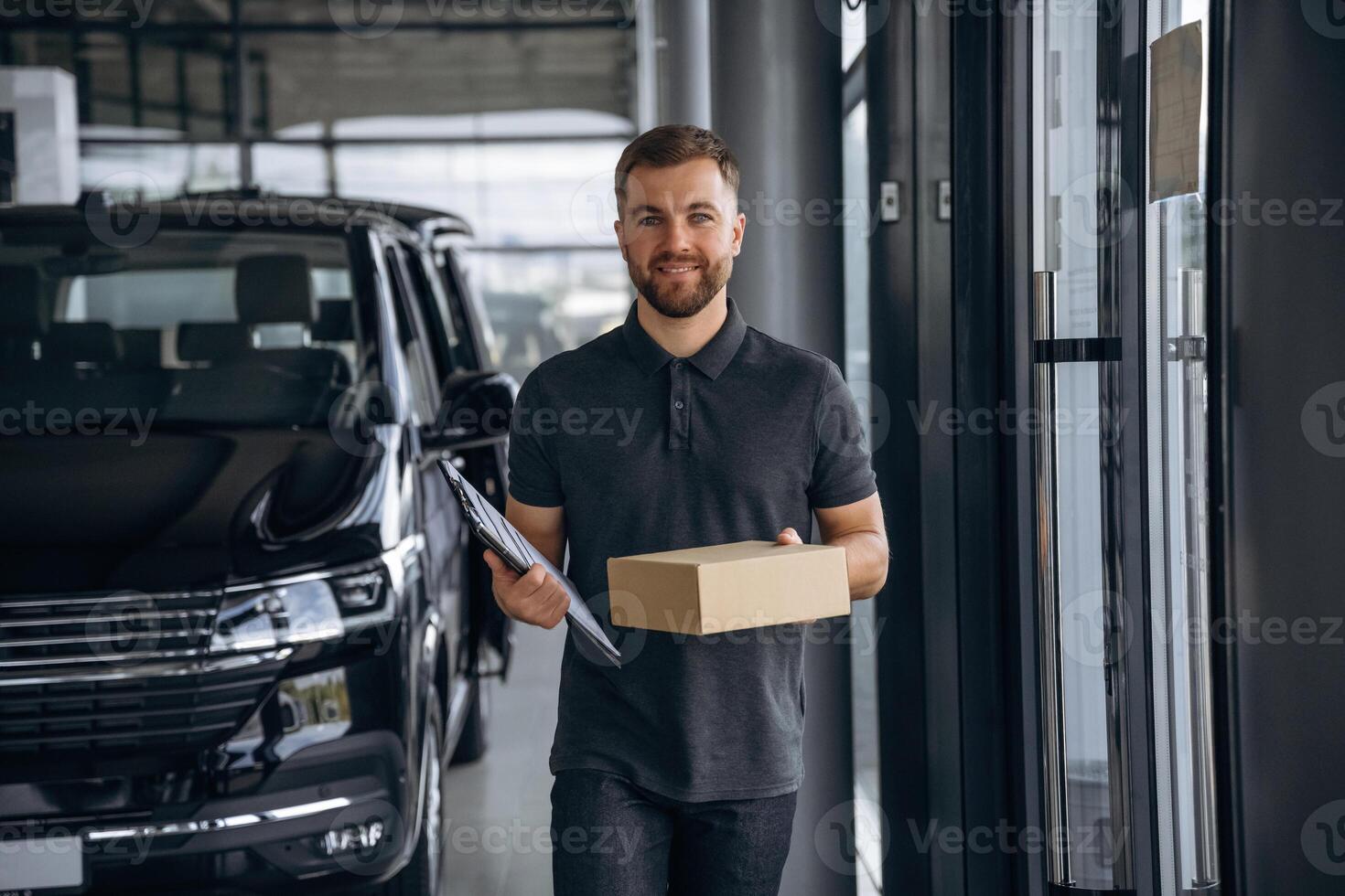 Standing,, holding notepad. Delivery ,man or loader is near the car with a box photo
