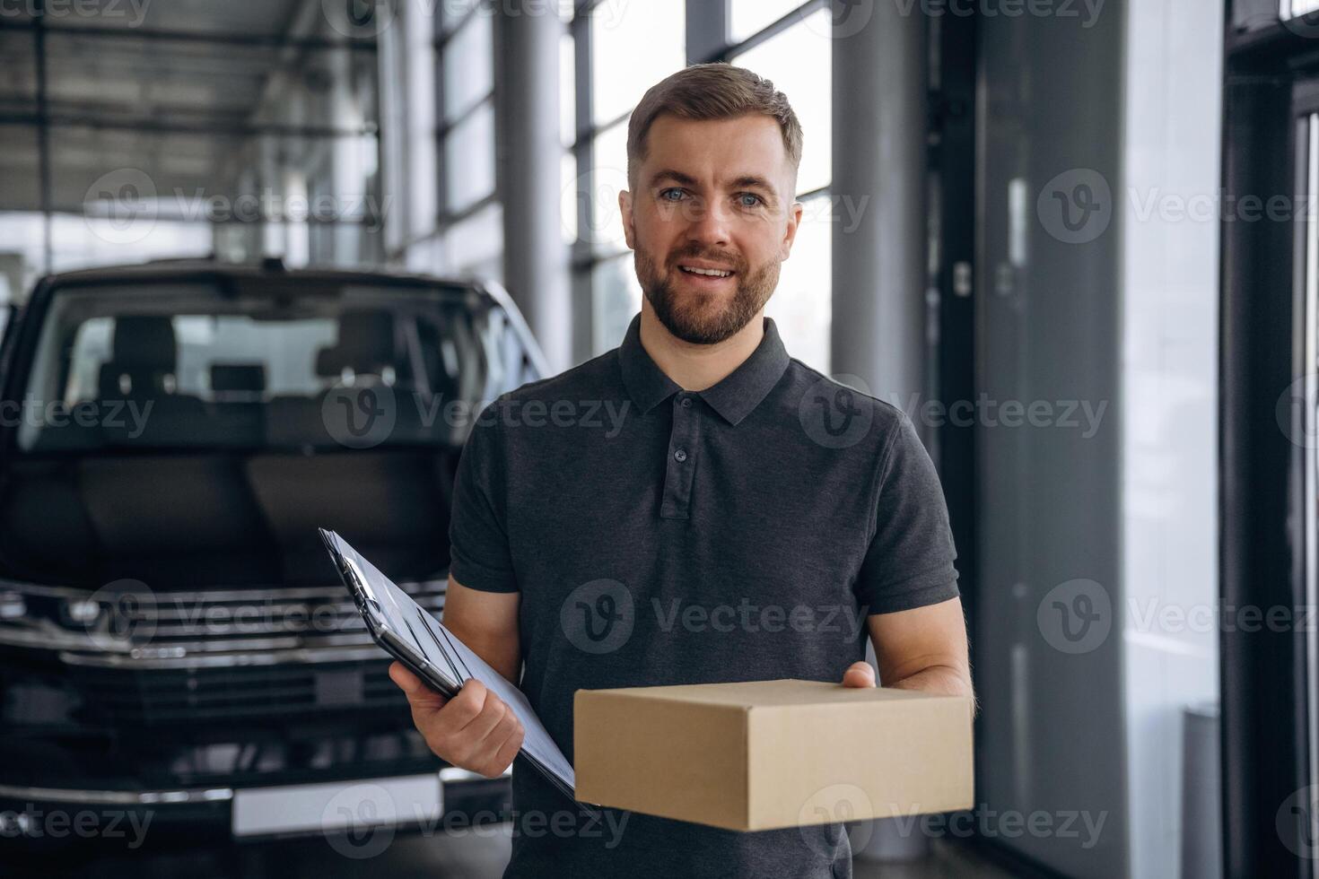 Standing, holding ,,,notepad. Delivery man or loader is near the car with a box photo