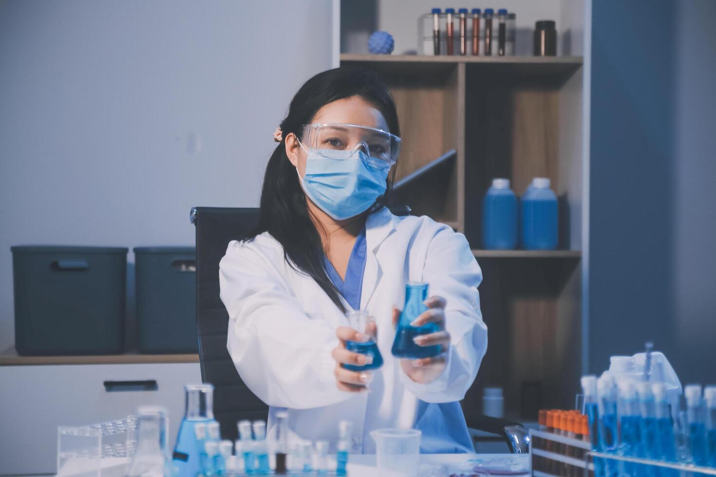 A woman in a lab coat and mask is holding a vial photo