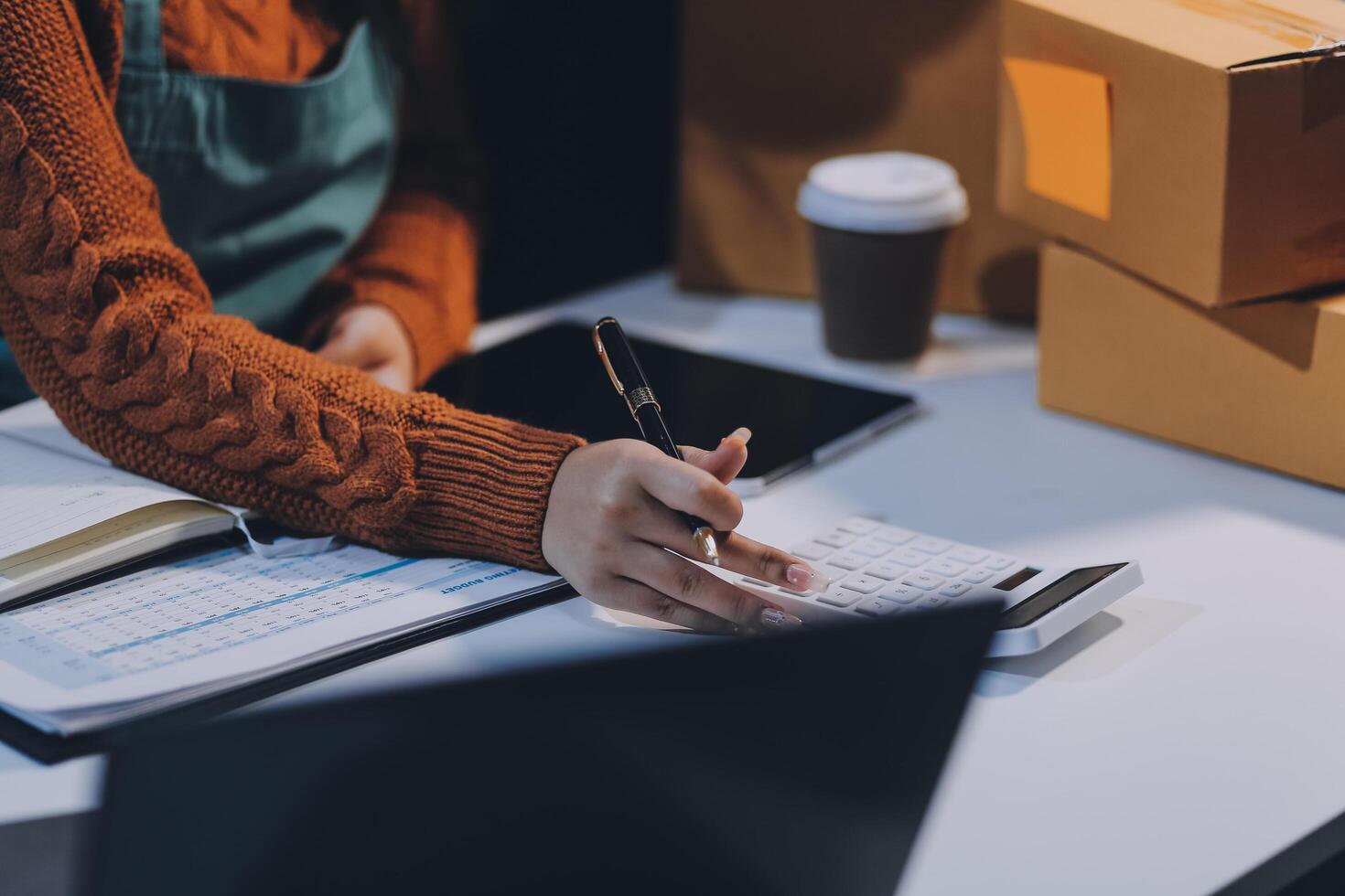 A woman is working on her laptop with a pen and paper in front of a desk with boxes photo