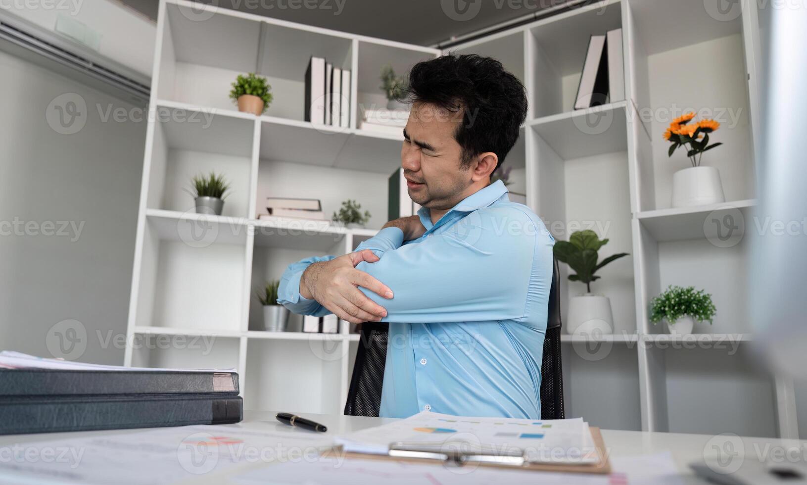 Young Businessman in Modern Office Experiencing Shoulder Pain While Working at Desk Surrounded by Documents and Shelves photo
