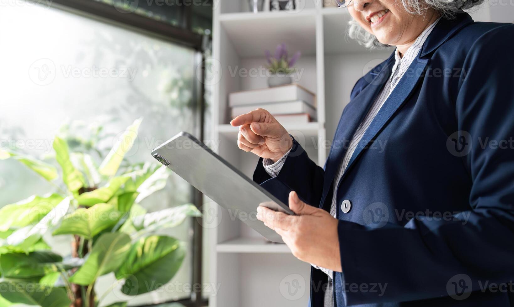 Elderly business woman reviewing documents on a tablet with a smile in a bright, green-filled office photo