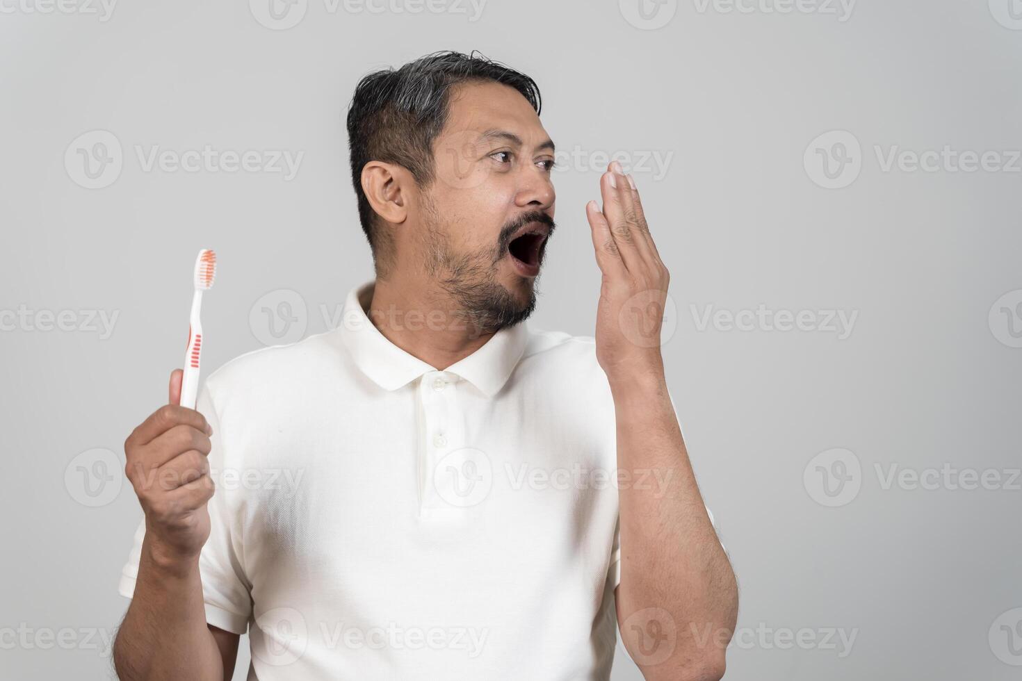 Asian man holding toothbrush and checking bad breath with hand covering mouth isolated on gray background photo