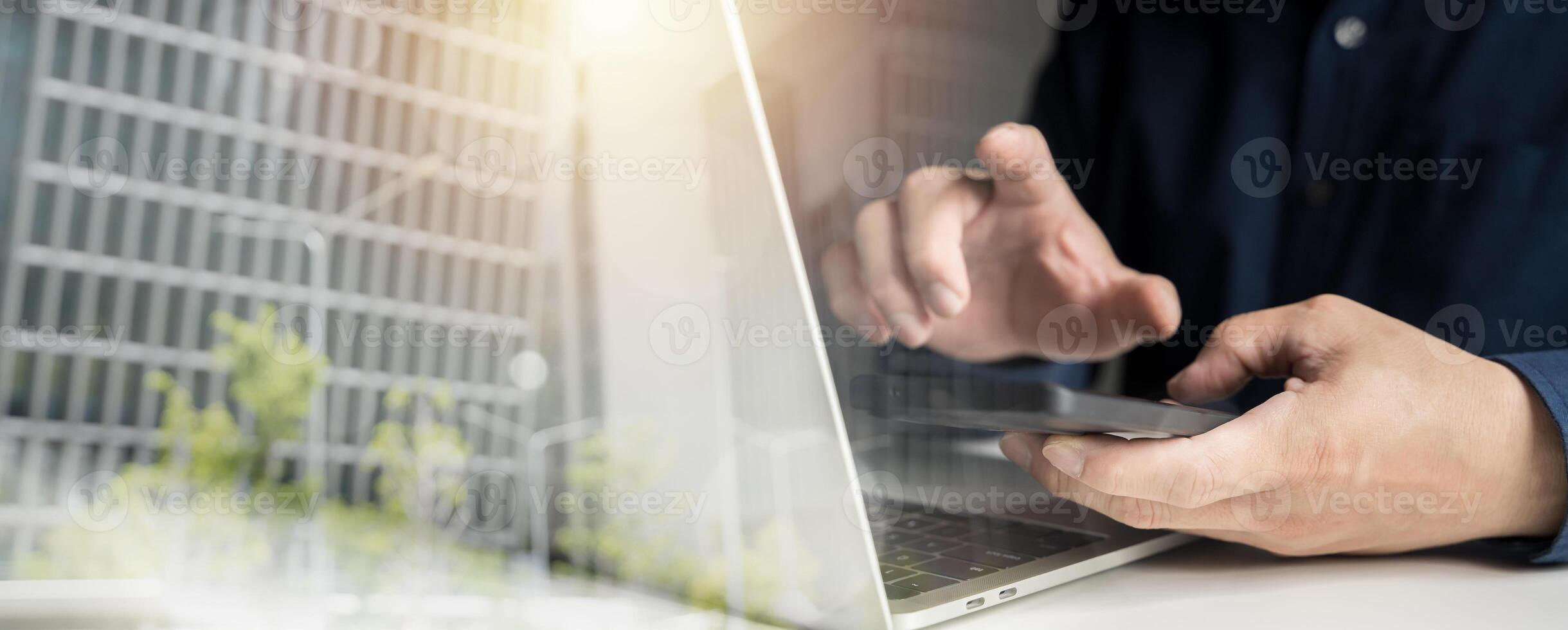 Businessman using smartphone and laptop computer in office with double exposure of glass building photo