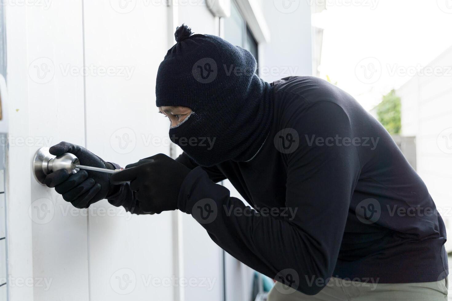 Masked burglar in black clothes using screwdriver to break into house through door lock during daylight robbery photo
