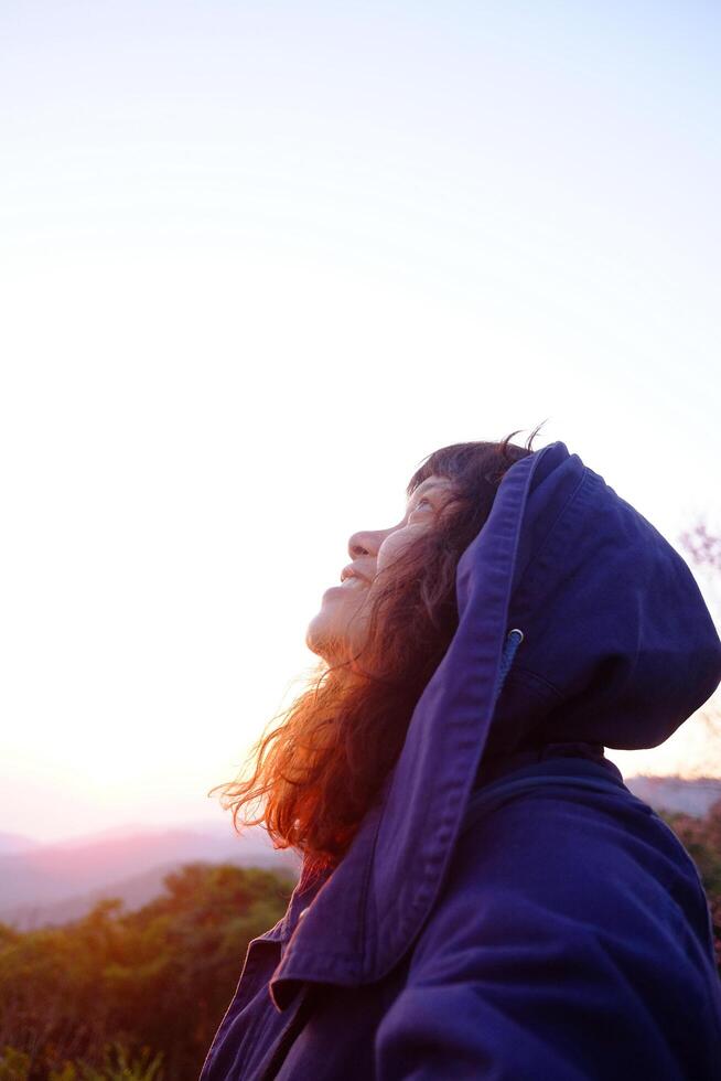 Traveler Asian woman wearing jacket and hood smiling in blooming wildflower on green lawn field with blue sky in winter on hill in Chiangmai of Thailand photo