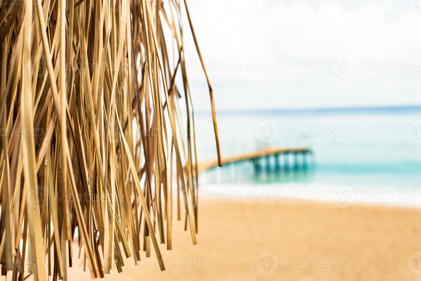 Thatched roof with tropical beach with sand and wooden pier on the sea shore in background. photo