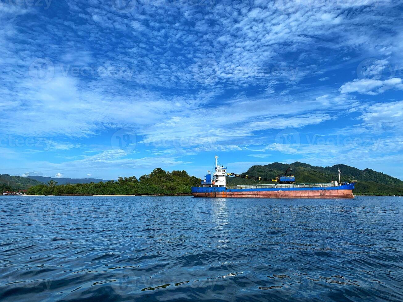 A cargo ship rests calmly near lush green hills under a vibrant sky, with clear blue water reflecting the peaceful tropical scene. photo