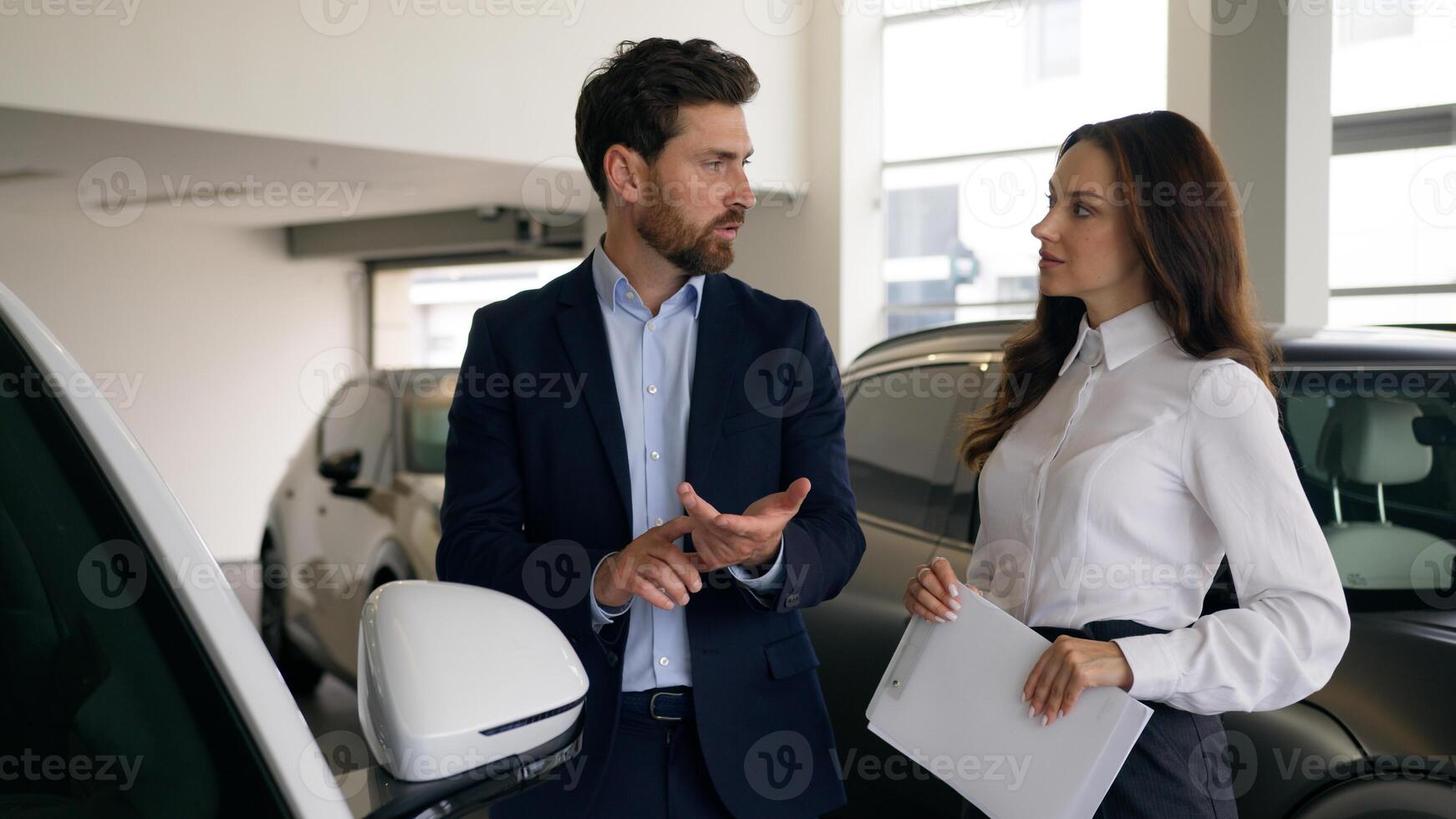 Two people talking in automobile showroom Caucasian man client businessman male choosing new luxury auto talking with woman saleswoman auto trader businessman buying new car with dealer communicate photo
