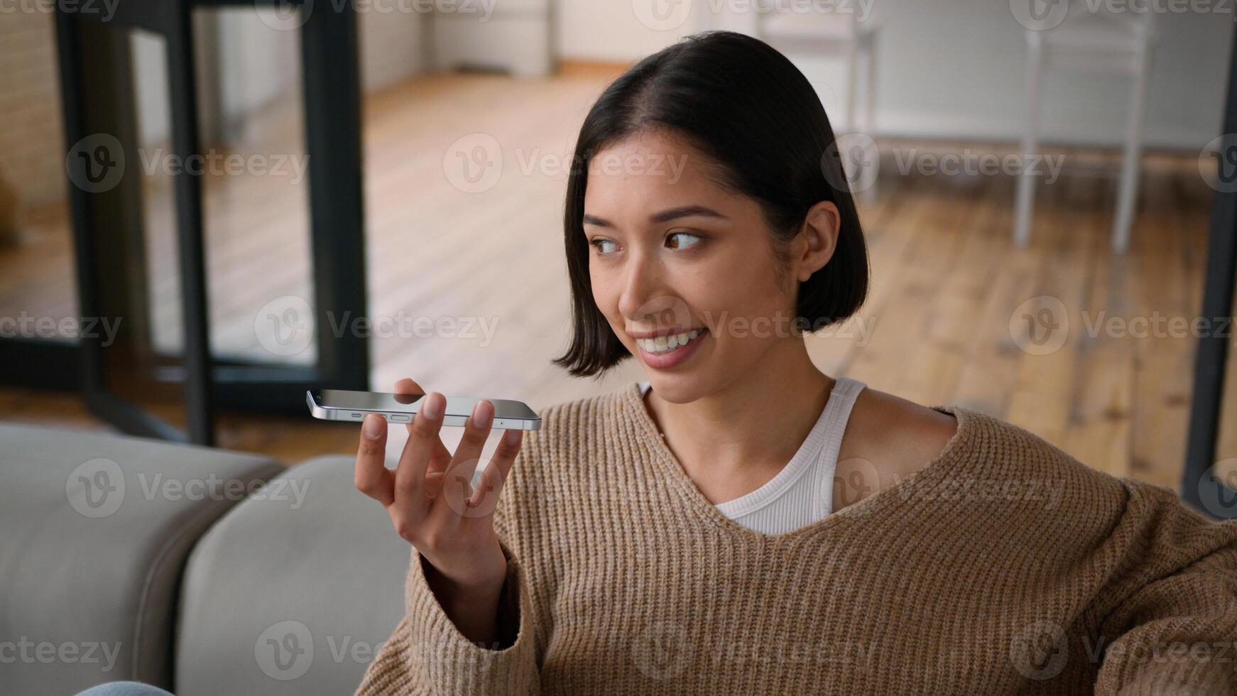 Happy joyful smiling Caucasian hispanic woman girl homeowner lady female user relax on home couch record audio message smartphone voice mail talking mobile phone speakerphone talk speak gossip laugh photo