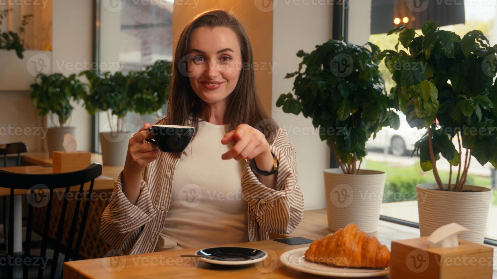 Caucasian woman sitting in cafe female girl drinking tea coffee hey you pointing gesture come here invitation looking at camera sit at table in cafeteria with croissant inviting welcome breakfast photo