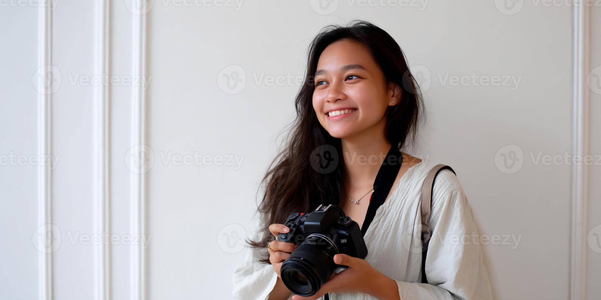 Young woman holding a camera, smiling, in front of white wall photo