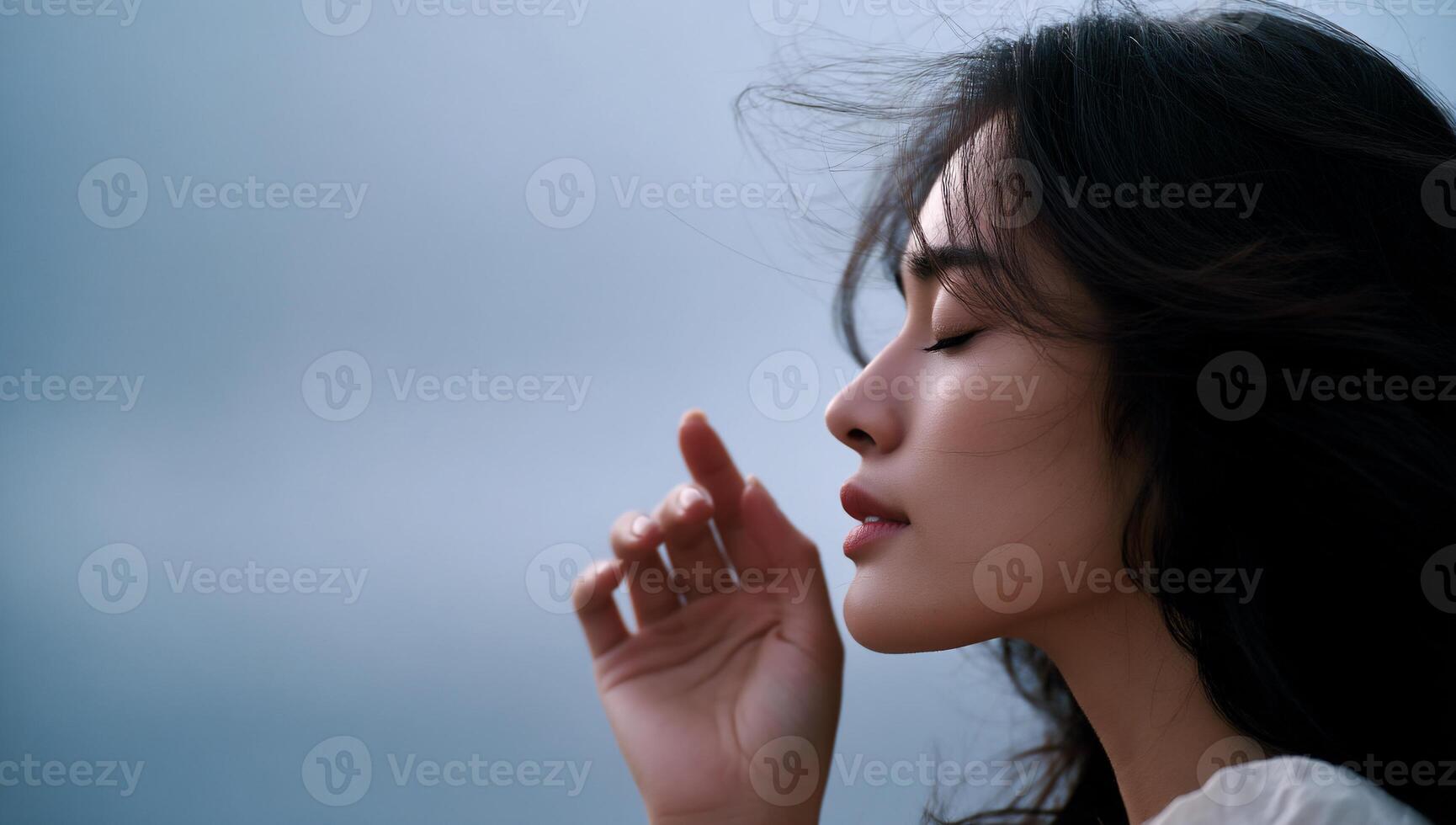 Woman's profile, eyes closed, touching finger to cheek, windswept hair against a soft, muted sky photo