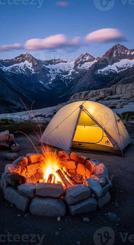 Cozy camping scene with campfire and tent under snowy mountain peaks at dusk photo