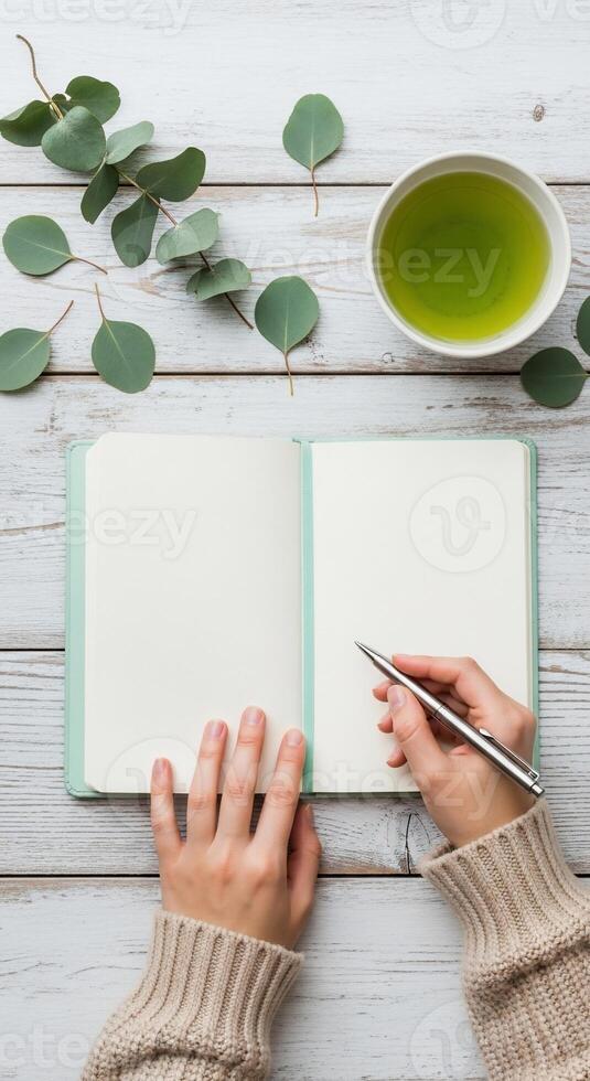 Top-down view of hands writing in a notebook with tea and eucalyptus leaves photo
