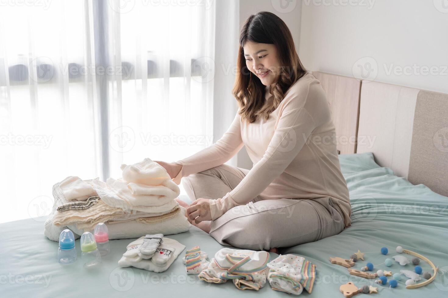 a Pregnant woman sitting on a bed with her baby's clothes photo