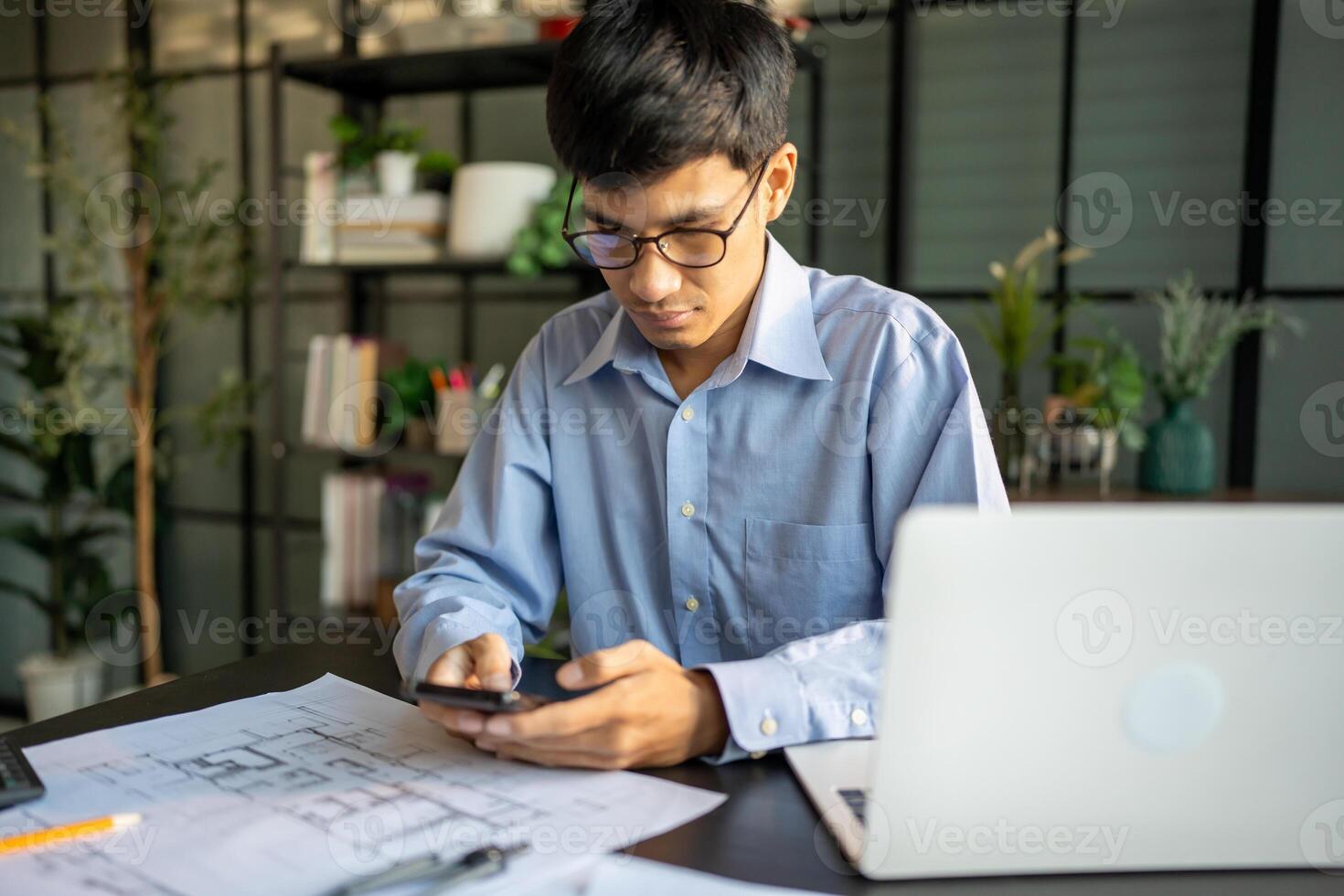 young man in glasses using his phone while working on blueprints photo