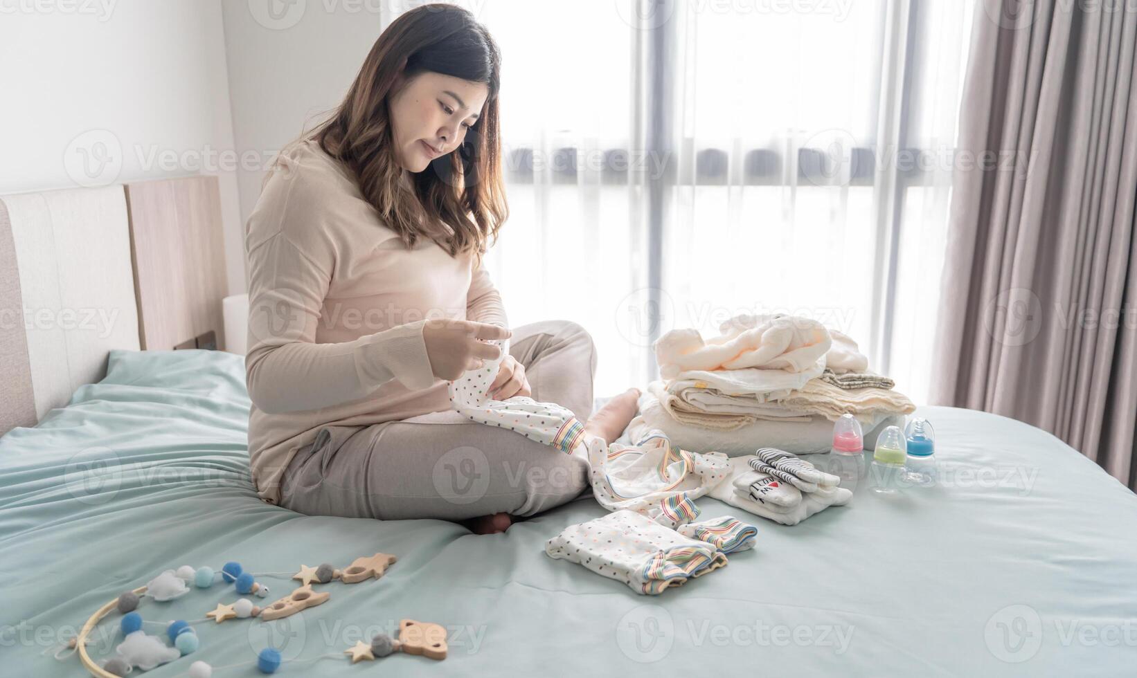 Pregnant asian woman getting ready for the maternity hospital preparing and planning baby clothes for new baby of pregnancy packing baby stuff on bed photo