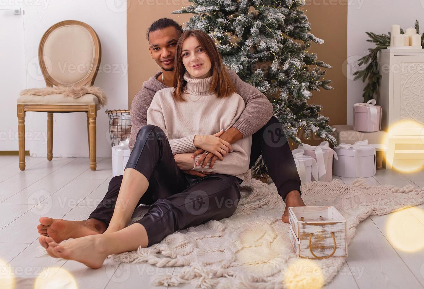 A happy stylish guy in a brown sweatshirt is sitting hugging his girlfriend, near the Christmas tree photo