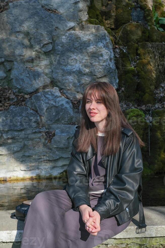 Young woman sitting by a stone wall near water on a sunny day in early spring photo