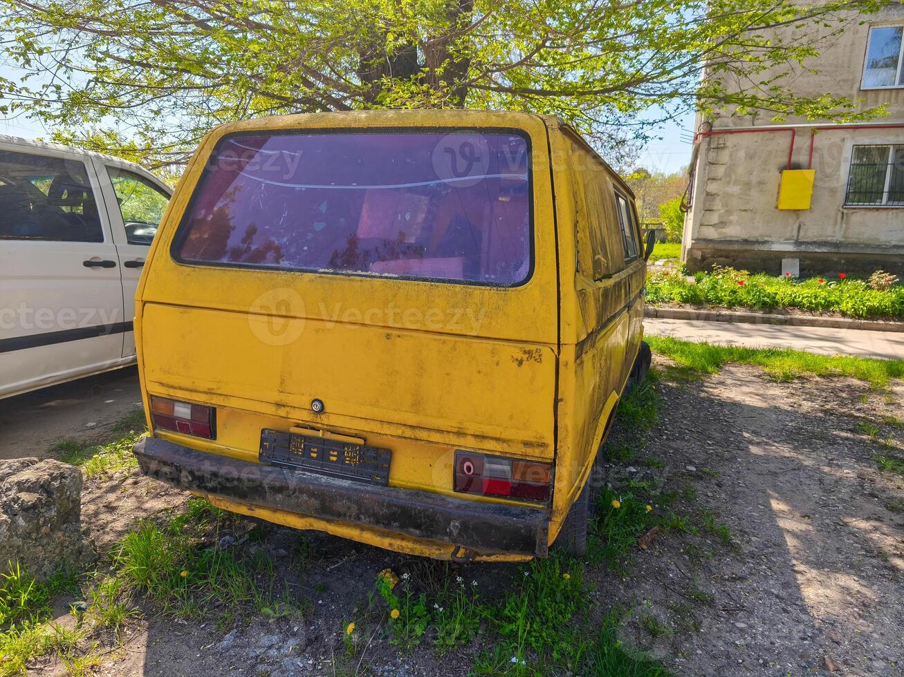 A yellow van is parked in a grassy area. The van is old and dirty, and it is parked next to a building photo