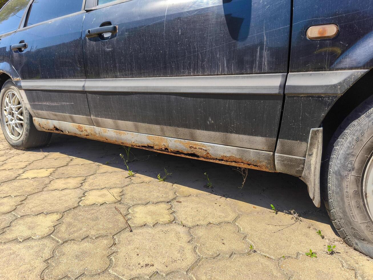 A dirty car with a tire on the ground. The car is parked on a brick road. The tire is flat and the car is in a state of disrepair photo