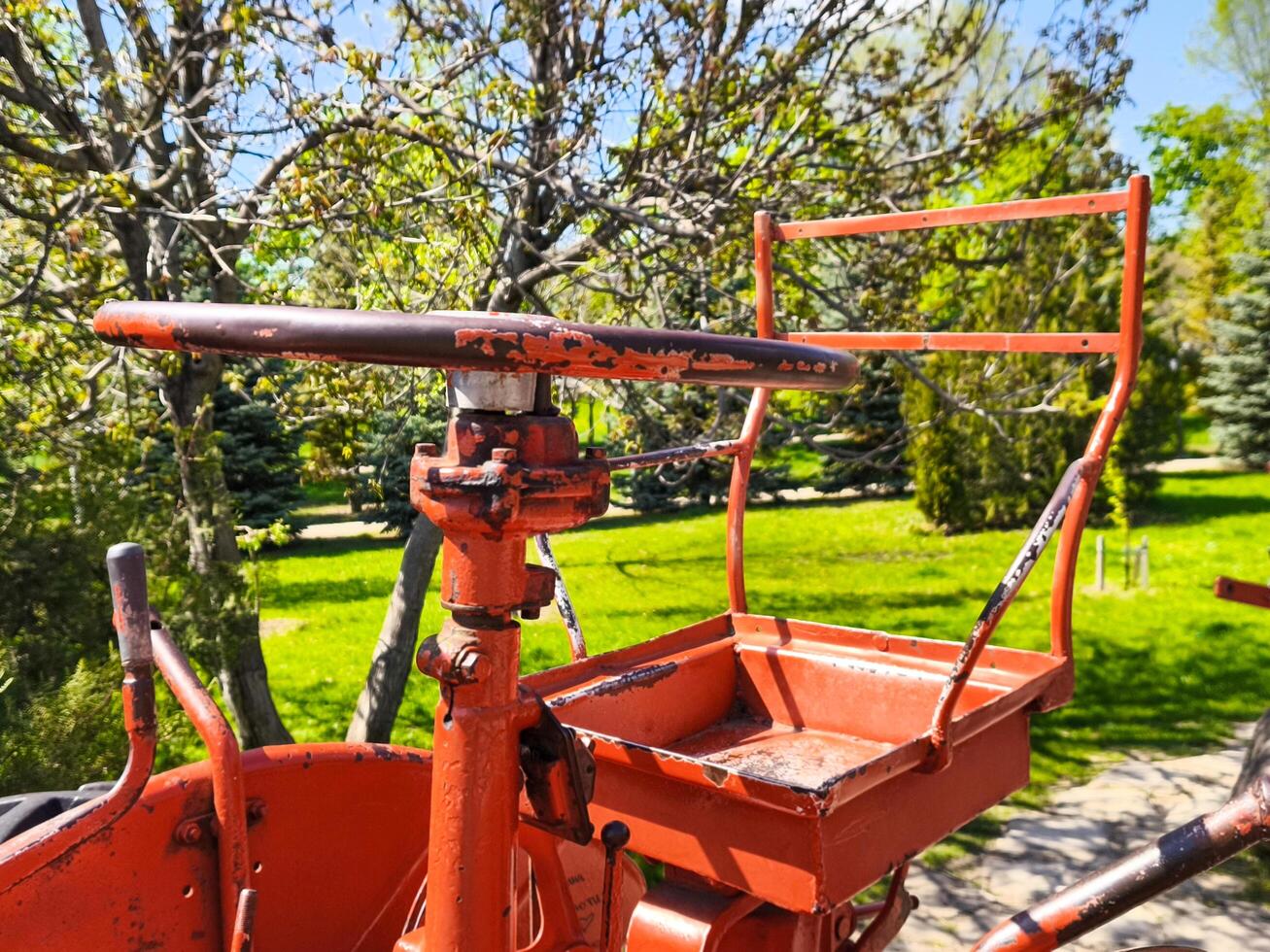 An old red machine with a rusty handle and a red tray. The machine is in a park with trees and grass photo