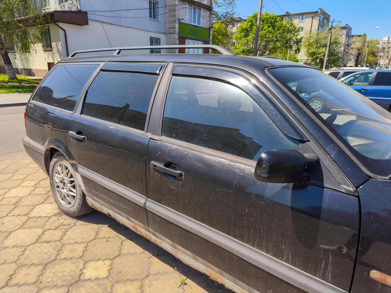 A black car with a dirty windshield sits in a parking lot. The car is parked next to a building with a white roof photo