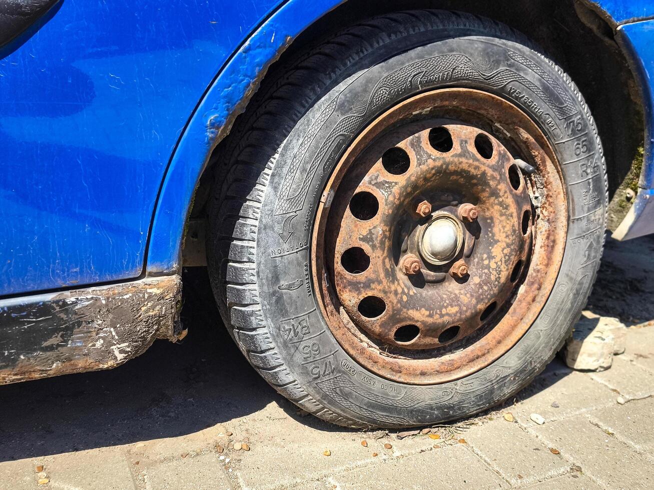 A rusted tire on a car. The tire is black and has a white rim. The tire is old and worn, and it is dirty. The car is parked on a street, and the tire is in the foreground photo