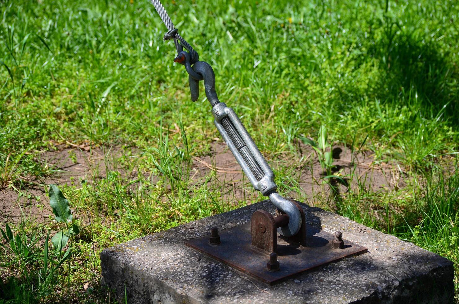A chain is attached to a metal object on a cement slab. The chain is attached to a rope, which is tied to a tree. The scene is peaceful and serene, with the grass photo
