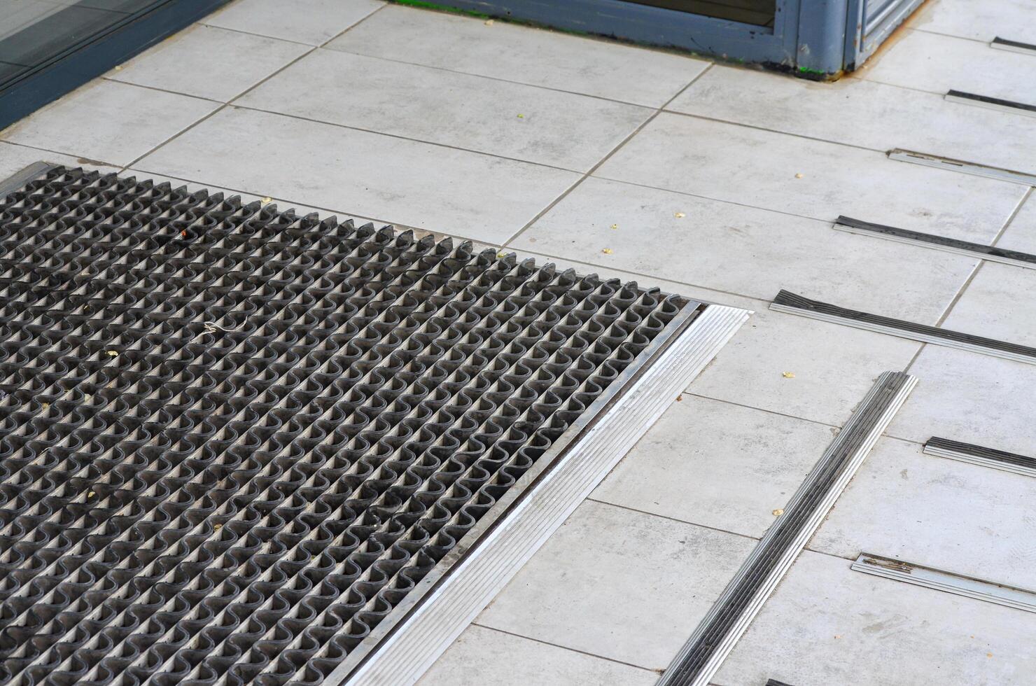 A metal grate with a black and silver design sits on a gray tile floor. The grate is positioned in front of a door, and the tile floor is tiled in a pattern. Concept of order and structure photo