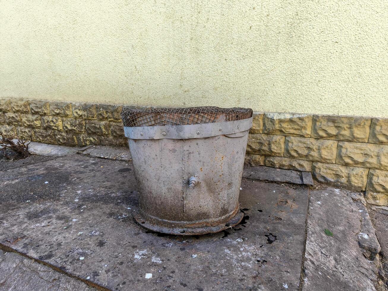 A dirty old bucket sits on the ground next to a wall. The bucket is old and worn, and it appears to have been used for a long time. The scene is somewhat bleak and uninviting photo