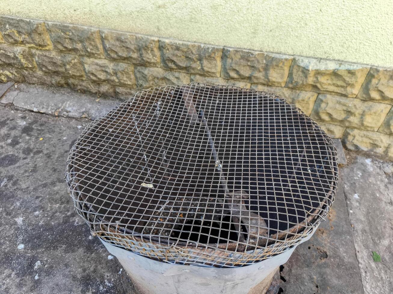 A metal trash can with a mesh lid is sitting on a sidewalk. The mesh lid is damaged and the trash can is currently empty photo