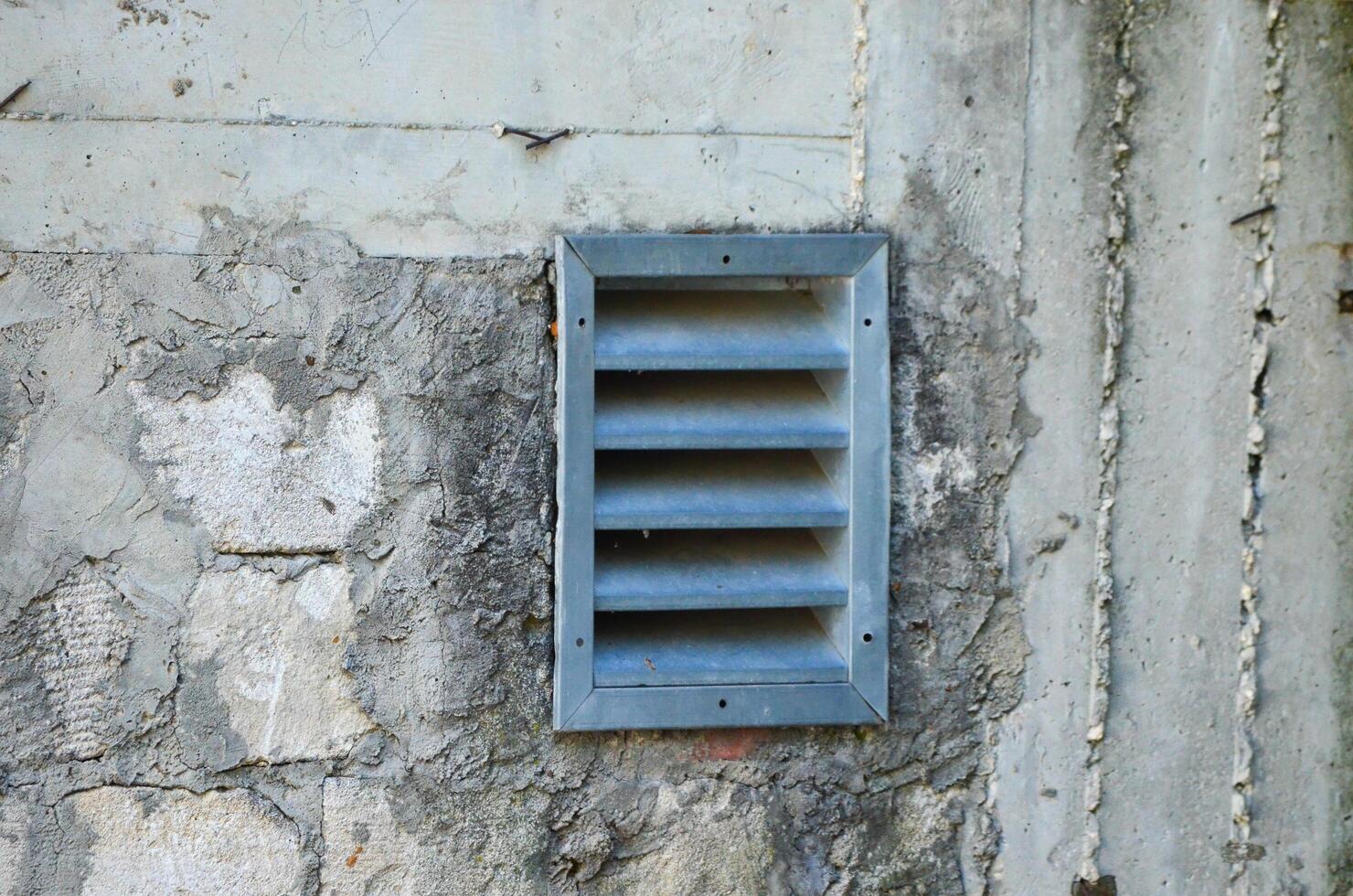 A vent is on a wall. The vent is open and has a metal grate. The vent is on a wall that is dirty and has some cracks photo