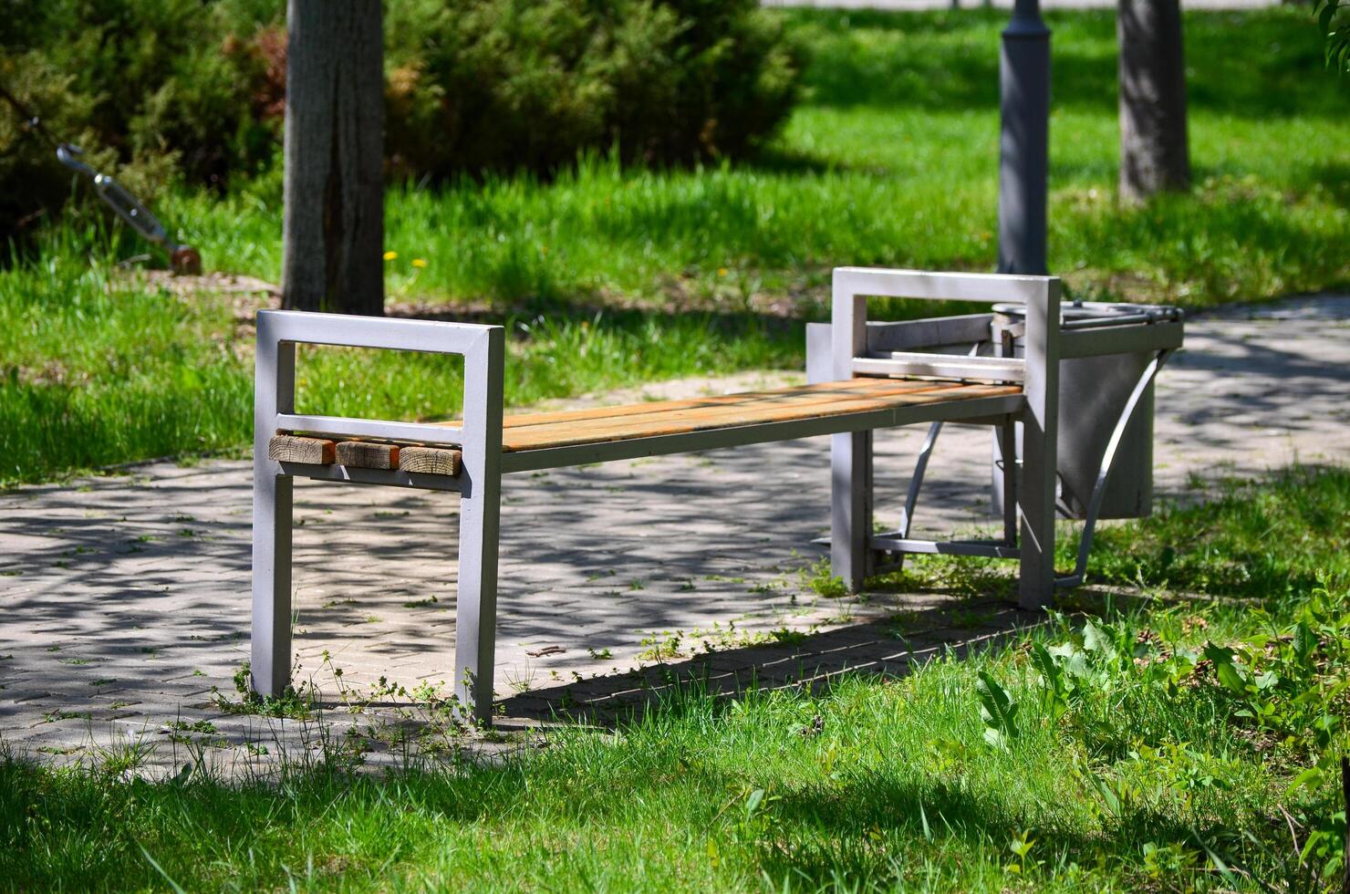 A park bench is sitting in the grass next to a trash can. The bench is empty and the grass is lush and green photo