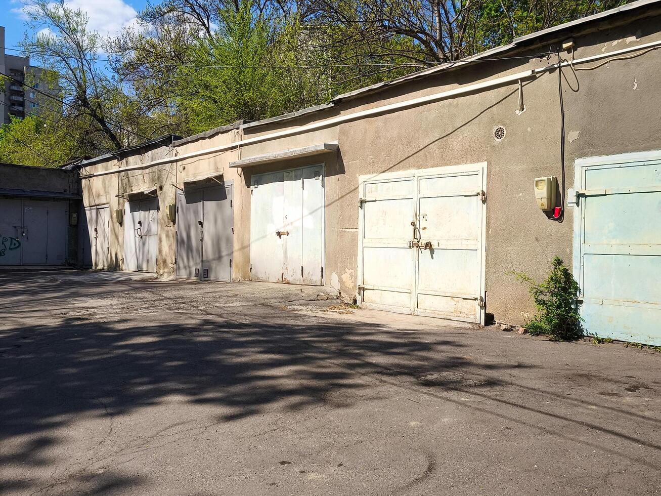 A row of old, abandoned garages sit empty in a parking lot. The buildings are grey and the sky is blue photo
