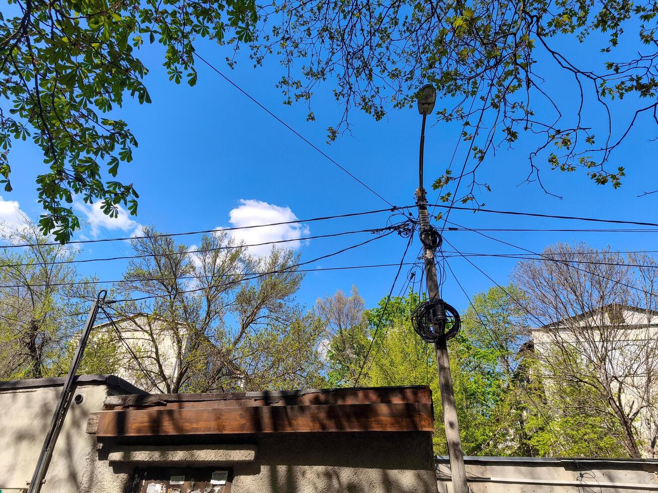 A tall pole with wires and a street light on top. The sky is blue and there are trees in the background photo
