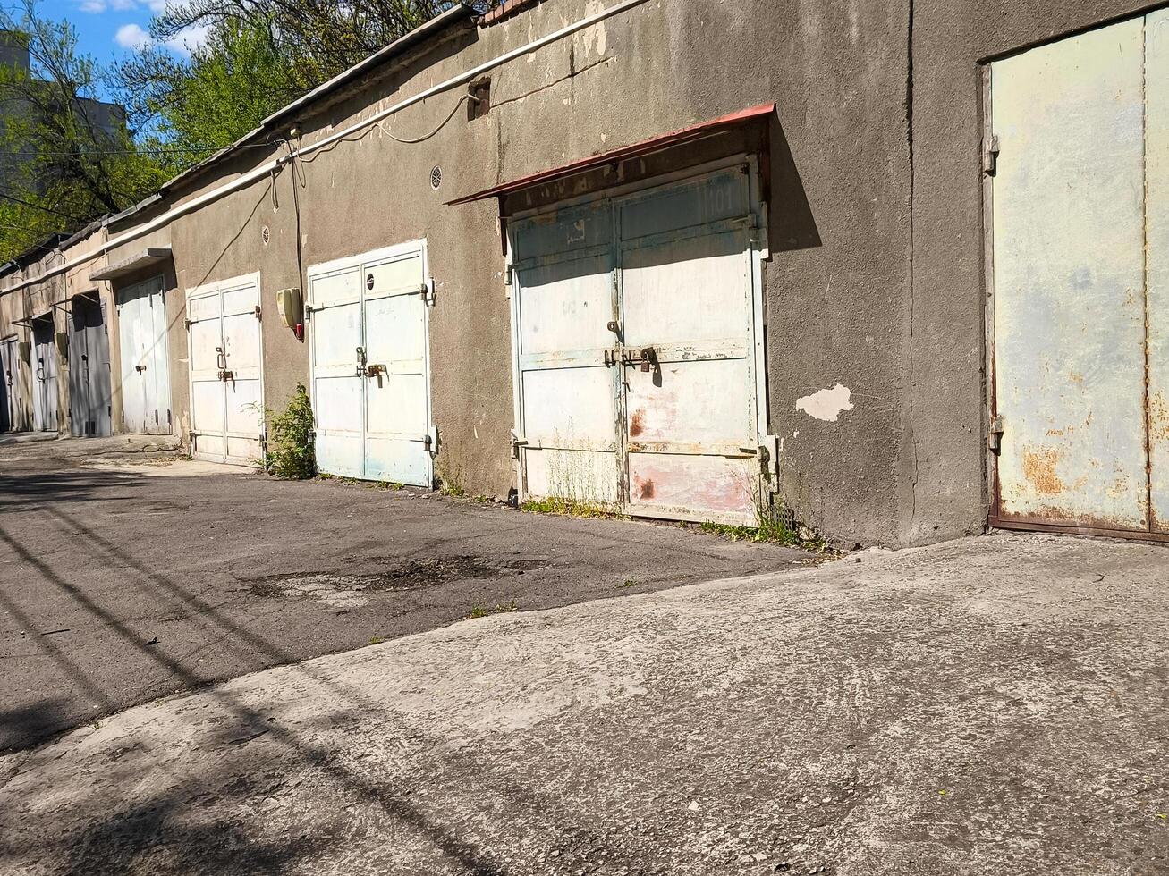 A row of old, abandoned garages sit empty on a street. The buildings are grey and the sky is blue photo