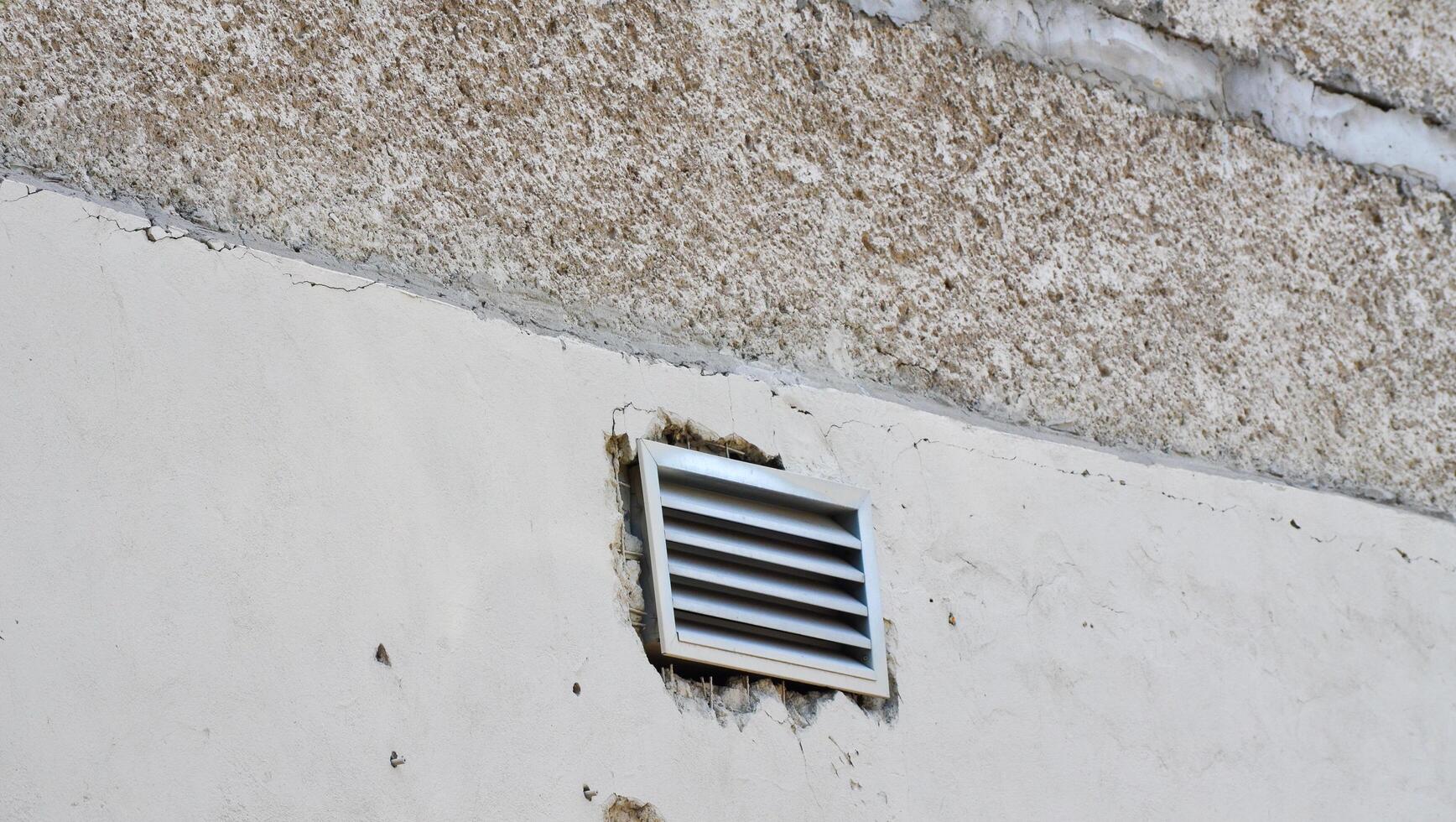 A vent is on the wall of a building. The vent is white and has a black grate photo