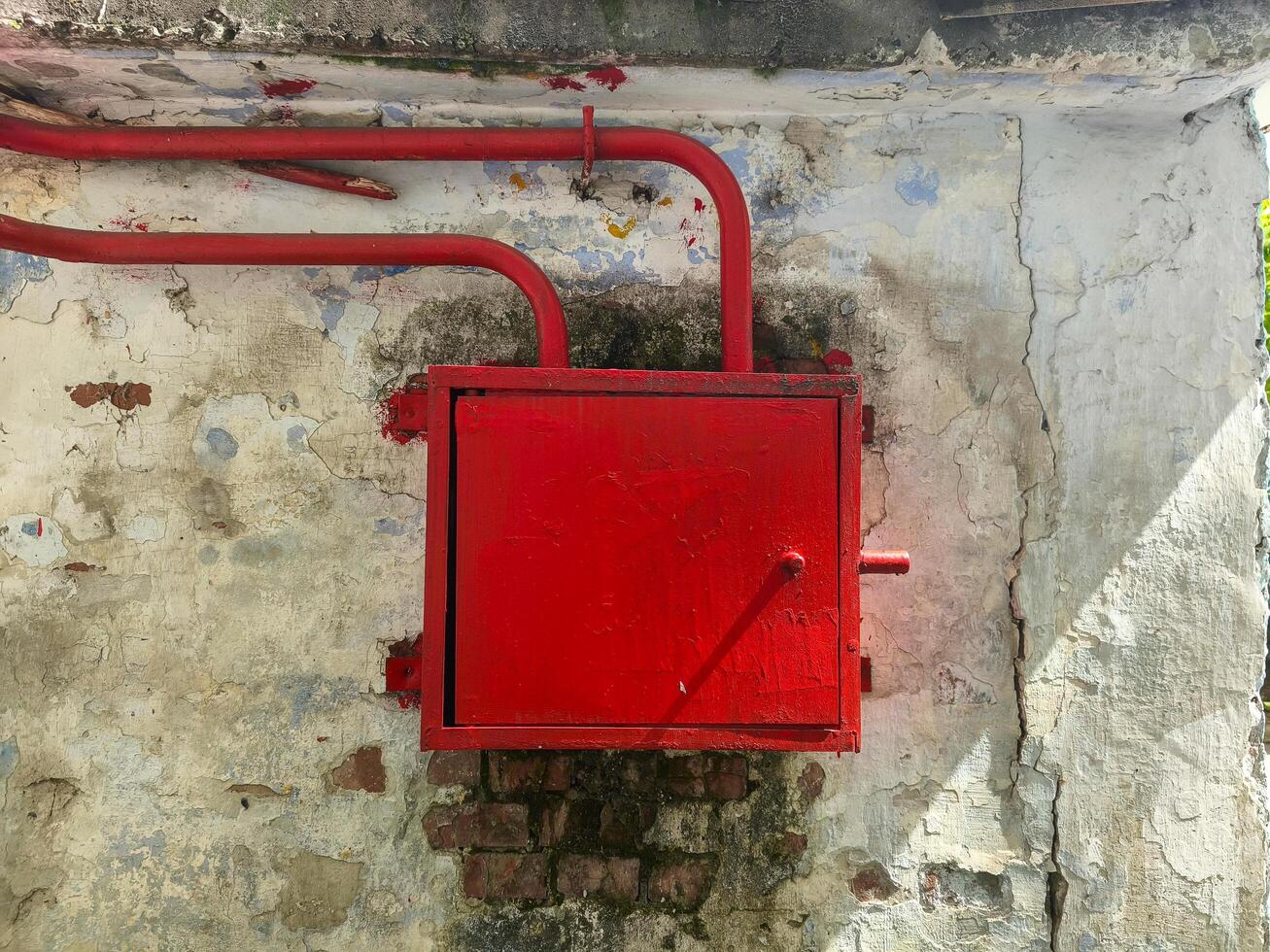 A red box with a red handle is sitting on a wall. The box is made of metal and has a red handle. It is located on a wall that is made of bricks and has a white color photo