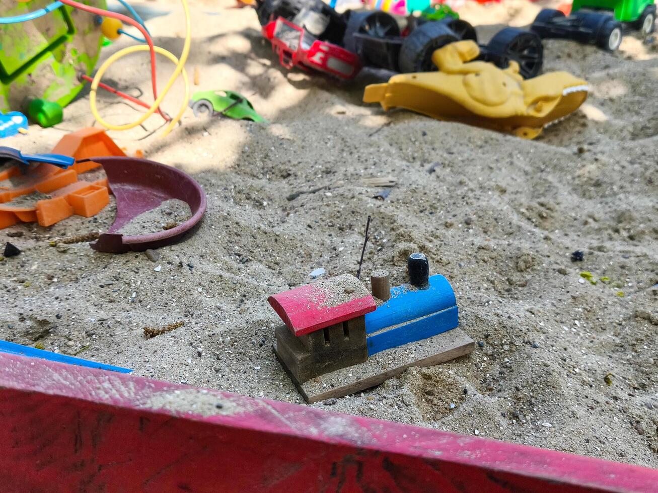 A toy train is sitting in the sand next to a pile of toys. The scene is messy and disorganized, with toys scattered all over the ground. The train is the only item that seems to be in order photo