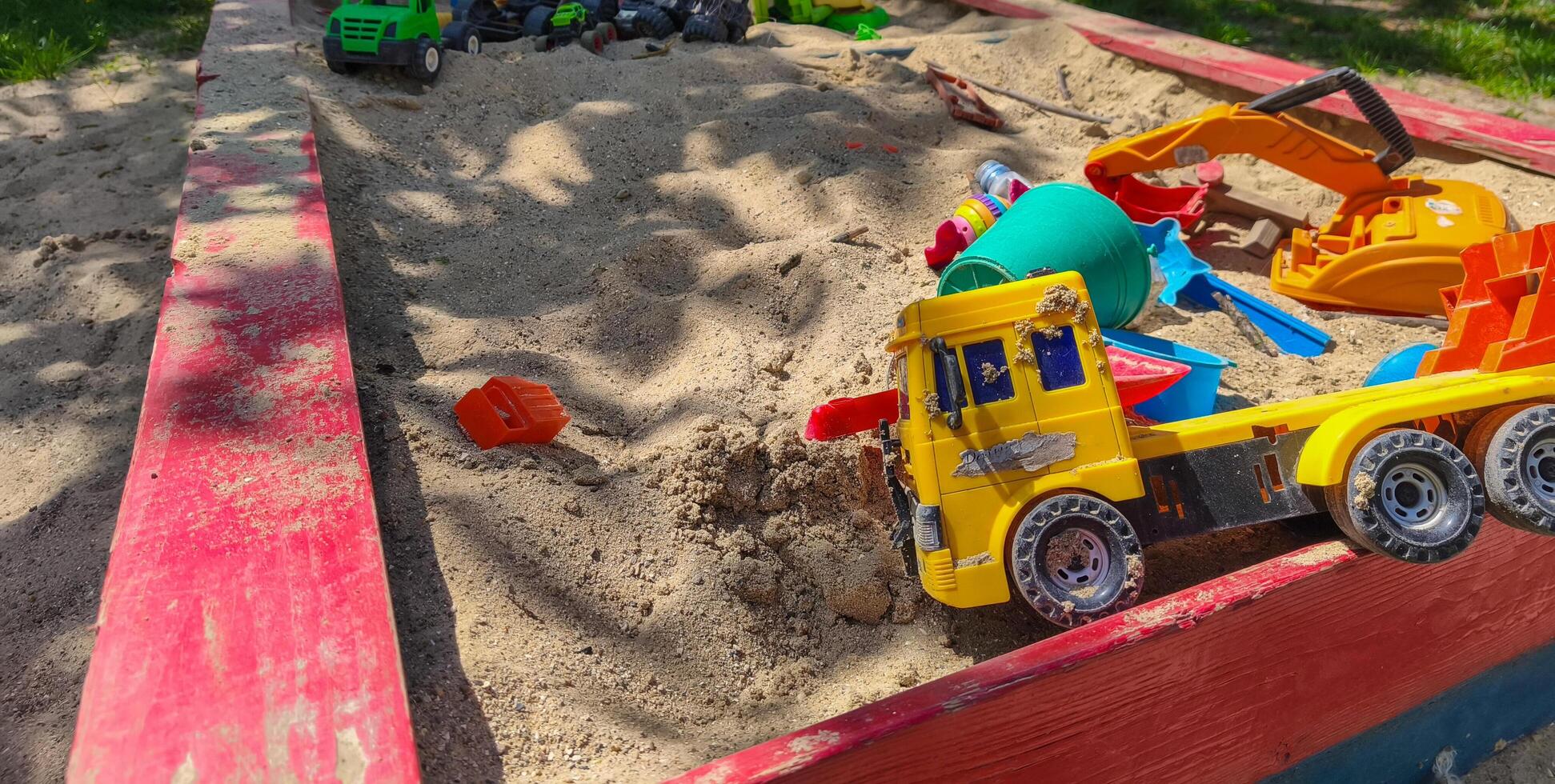 A toy truck is sitting in a sandbox with other toys. The sandbox is red and has a wooden border photo