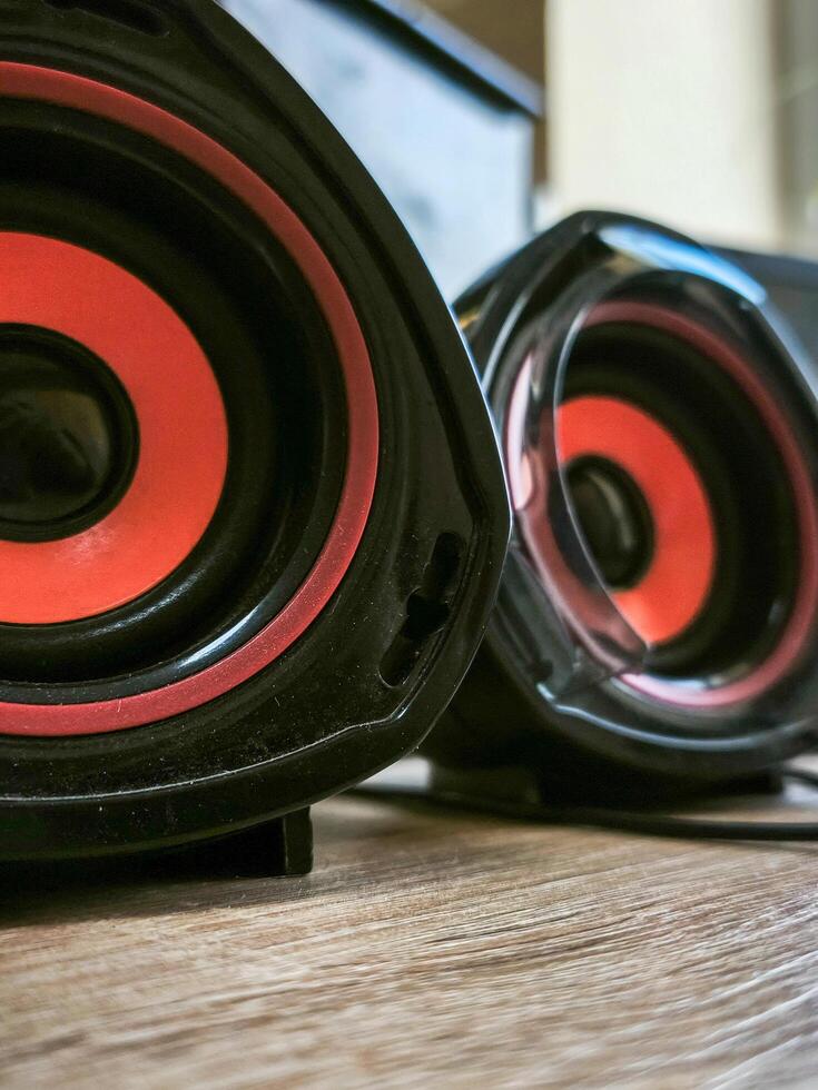Two speakers with red and black accents sit on a wooden table. The speakers are black and red, and they are placed on a wooden surface photo