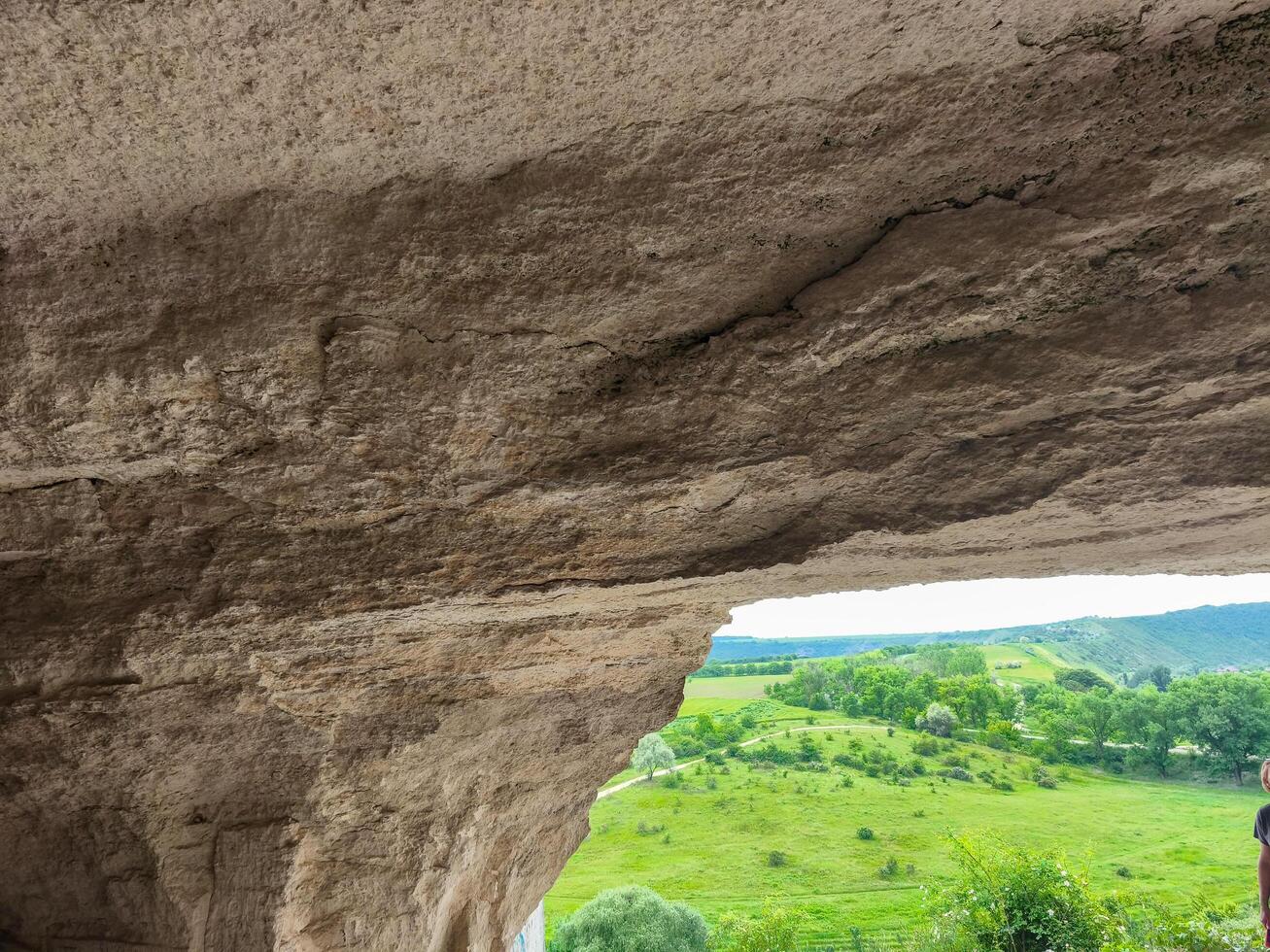 A large rock formation with a small opening in the middle. The opening is above a grassy field photo