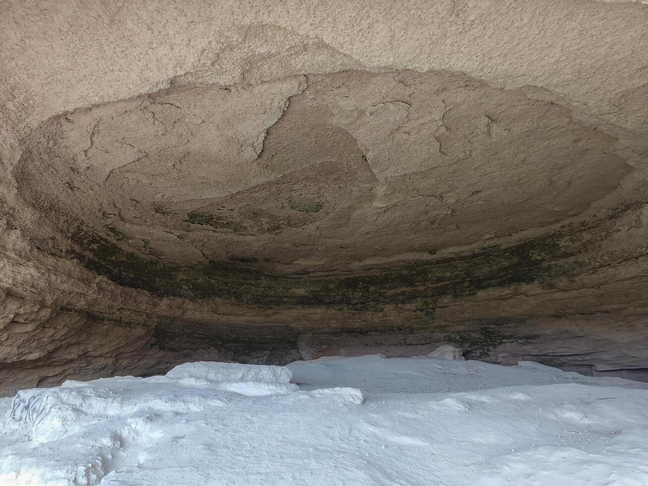 A cave with a large hole in the ceiling. The ceiling is covered in dirt and the floor is covered in white photo